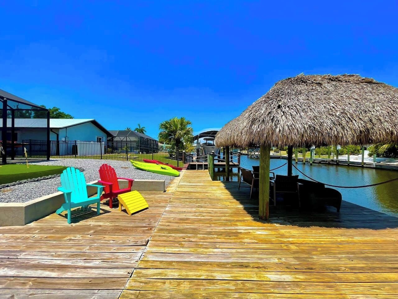 A wooden pier leading to a thatched-roof structure over water, with colorful Adirondack chairs and a small dock.