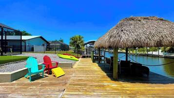 A wooden pier leading to a thatched-roof structure over water, with colorful Adirondack chairs and a small dock.
