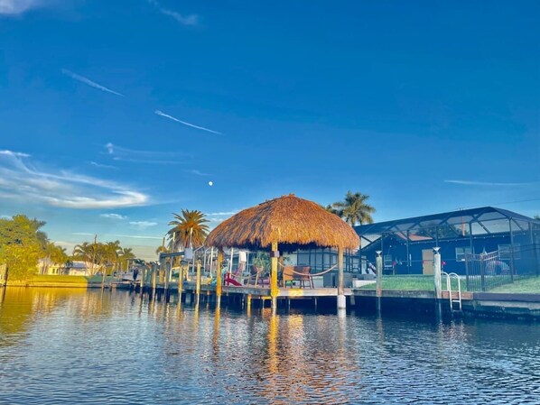 A thatched-roof structure with a pool and lounge chairs, surrounded by water and palm trees.