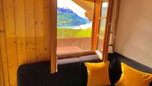 Um quarto aconchegante com teto de madeira, uma janela grande com vista para uma paisagem montanhosa e um sofá escuro com duas almofadas amarelas.