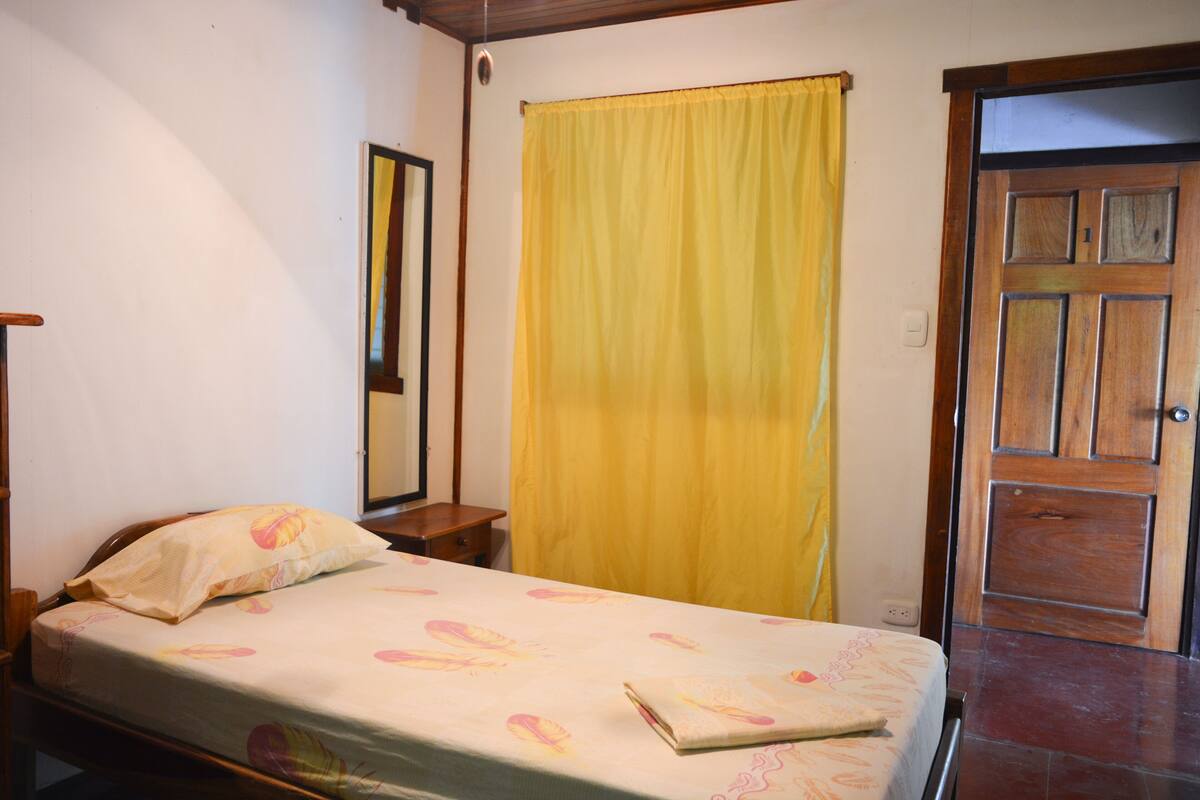 A bedroom with a bed, a wooden door, a yellow curtain, and a mirror on the wall.