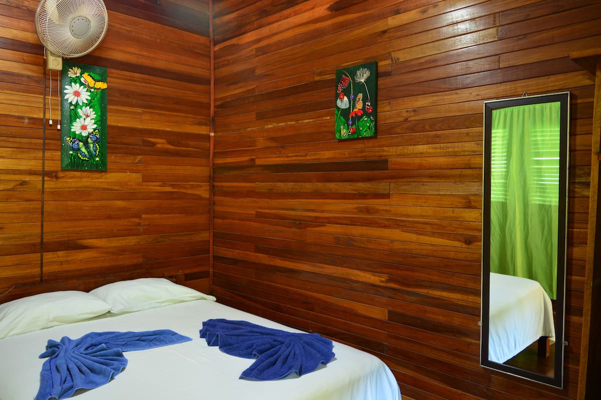 A wooden-paneled room with a bed, a fan, and two paintings on the wall.