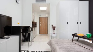 A modern hotel room with a bed, a desk with a chair, a TV, a wardrobe, and a small table with a cup and a pastry. - Modern Studio | Parking | Air Conditioning | Close to Expo XXI (Warszawa)