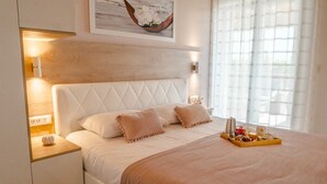 Ein ordentlich eingerichtetes Hotelzimmer mit einem großen Bett, einem Nachttisch mit Lampe, einem Tablett mit Frühstücksgebäck und einem gerahmten Kunstwerk über dem Bett.