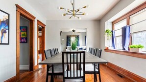 A dining area with a long table, multiple chairs, a modern chandelier, and a window with curtains.