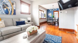 A modern living room with a grey sofa, a coffee table with a vase of flowers, a blue patterned rug, and a flat-screen TV mounted on the wall.