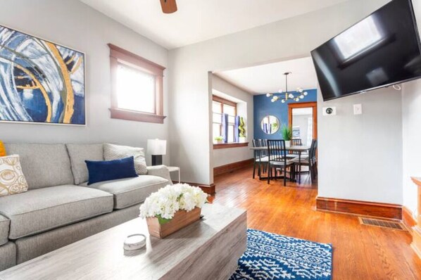Ein modernes Wohnzimmer mit einem grauen Sofa, einem Couchtisch mit Blumenvase, einem blau gemusterten Teppich und einem flachbildfernseher an der Wand.