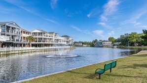 A waterfront property with a fountain, a grassy area with a bench, and a row of multi-story buildings.