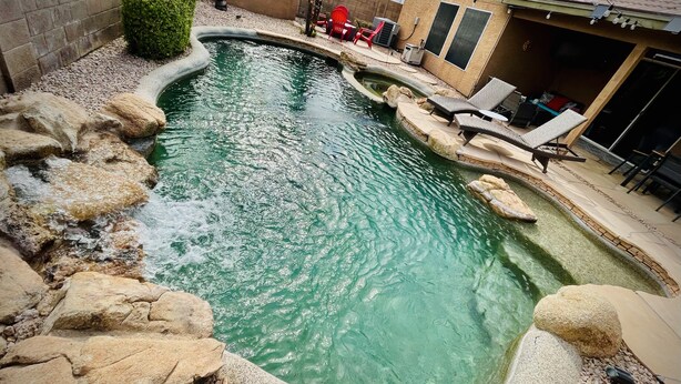 A swimming pool with a curved design, surrounded by rocks and pebbles. There are lounge chairs and a covered seating area in the background. - Casa Del Sol Gilbert, Arizona Heated Pool & Hot Tub (Gilbert)