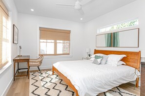 Ein Schlafzimmer mit einem hölzernen Bett, einem Schreibtisch, einem Stuhl, einem großen Fenster mit Jalousien und einem Deckenventilator.
