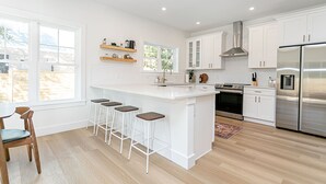 A modern kitchen with white cabinetry, a central island with bar stools, a stainless steel refrigerator, and a large window providing natural light. - Luxury heated pool & spa home, pool bar, steps to the beach & two master suites! (St. Augustine)