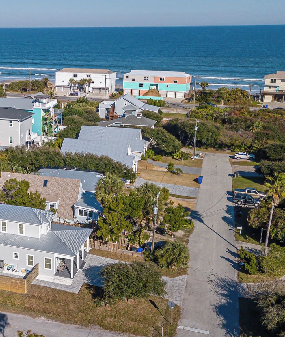An aerial view of a coastal residential area with houses, a road, and the ocean in the background.
