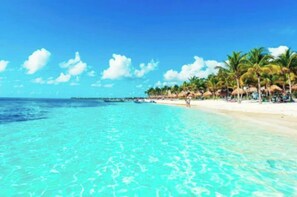 Ein Strand mit klarem türkisblauem Wasser, Palmen und einem in der Ferne liegenden Steg.