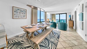 A modern living room with a wooden dining table, blue sofas, and a large window overlooking the ocean.