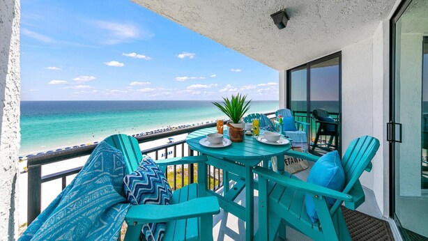 A balcony with turquoise furniture, a table with drinks and a plant, and a view of the ocean.