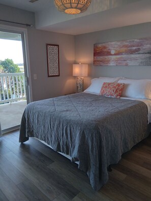 A bedroom with a large bed, a nightstand with a lamp, a painting on the wall, and a view through a sliding door. - Clean, Beachy, Amazing Waterview
Near Folly Beach & Downtown Charleston (Charleston)