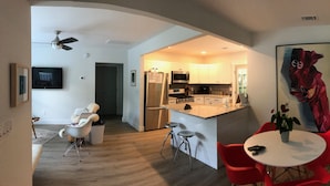 A modern kitchen with white cabinets, stainless steel appliances, and a central island. There is a dining area with red chairs and a white table. A large painting is on the wall, and a ceiling fan is visible. - Arthouse, freshly renovated 2 bedroom residential (North Charleston)