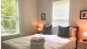 A neatly made bed with a white duvet and pillows, a stack of folded towels, and a nightstand with a lamp. - Arthouse, freshly renovated 2 bedroom residential (North Charleston)