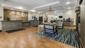 A spacious hotel lobby with a stone accent wall, a reception desk, a dining area with a patterned rug, and a kitchen in the background. - Best Western Plus Rumford Falls (Rumford)