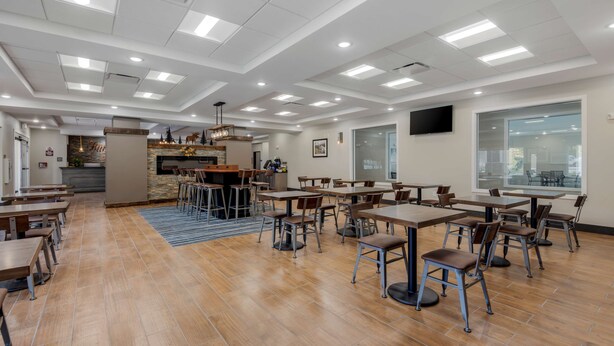 A spacious dining area with multiple tables and chairs, a kitchen area, and a large window providing natural light. - Best Western Plus Rumford Falls (Rumford)