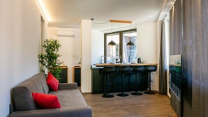 Ein modernes Wohnzimmer mit einem grauen Sofa, roten Kissen und einer großen Pflanze. Es gibt eine Küchenzeile mit Barhocker und Pendelleuchten.