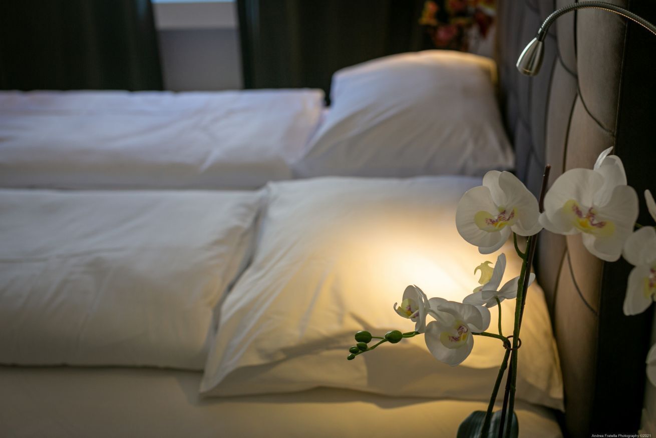 A neatly made bed with white linens and a single orchid in a vase.