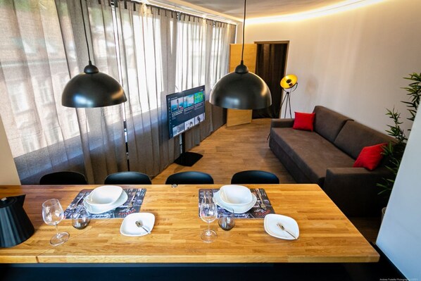 Una sala de estar moderna con una mesa de comedor de madera dispuesta para dos personas, un sofá con cojines rojos y un televisor montado en la pared.