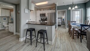 A modern kitchen with a central island, bar stools, and a dining area with wooden chairs. - PATHWAY TO THE SEA, 3RD FLOOR PENTHOUSE! HEATED POOL AND STEPS FROM OCEAN! (Corpus Christi)