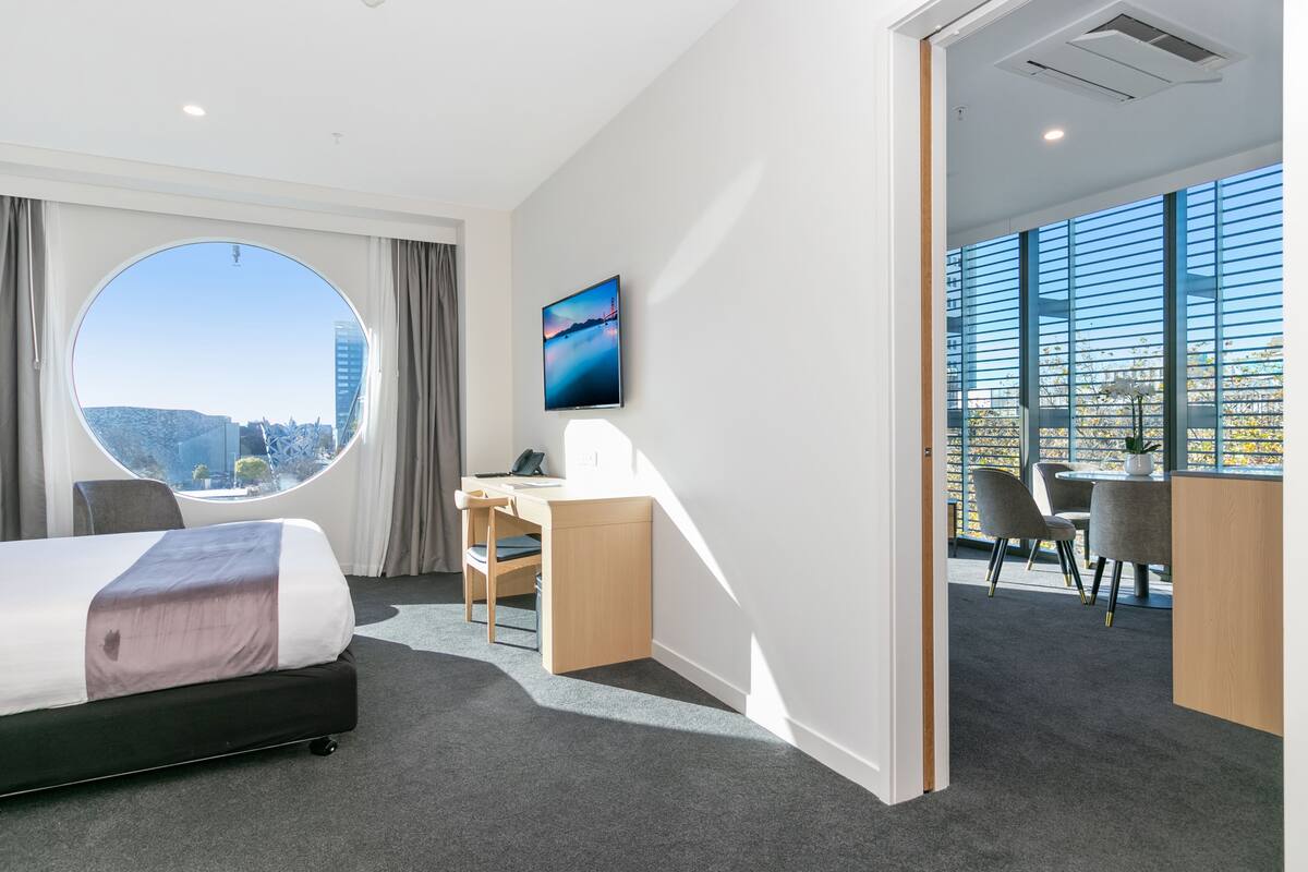 A modern hotel room with a bed, desk, and large windows offering a city view.