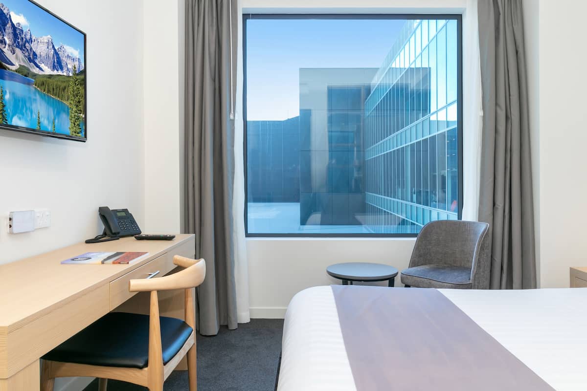 A hotel room with a bed, a desk with a chair, a small table, a phone, and a window with a view of a modern building and mountains.