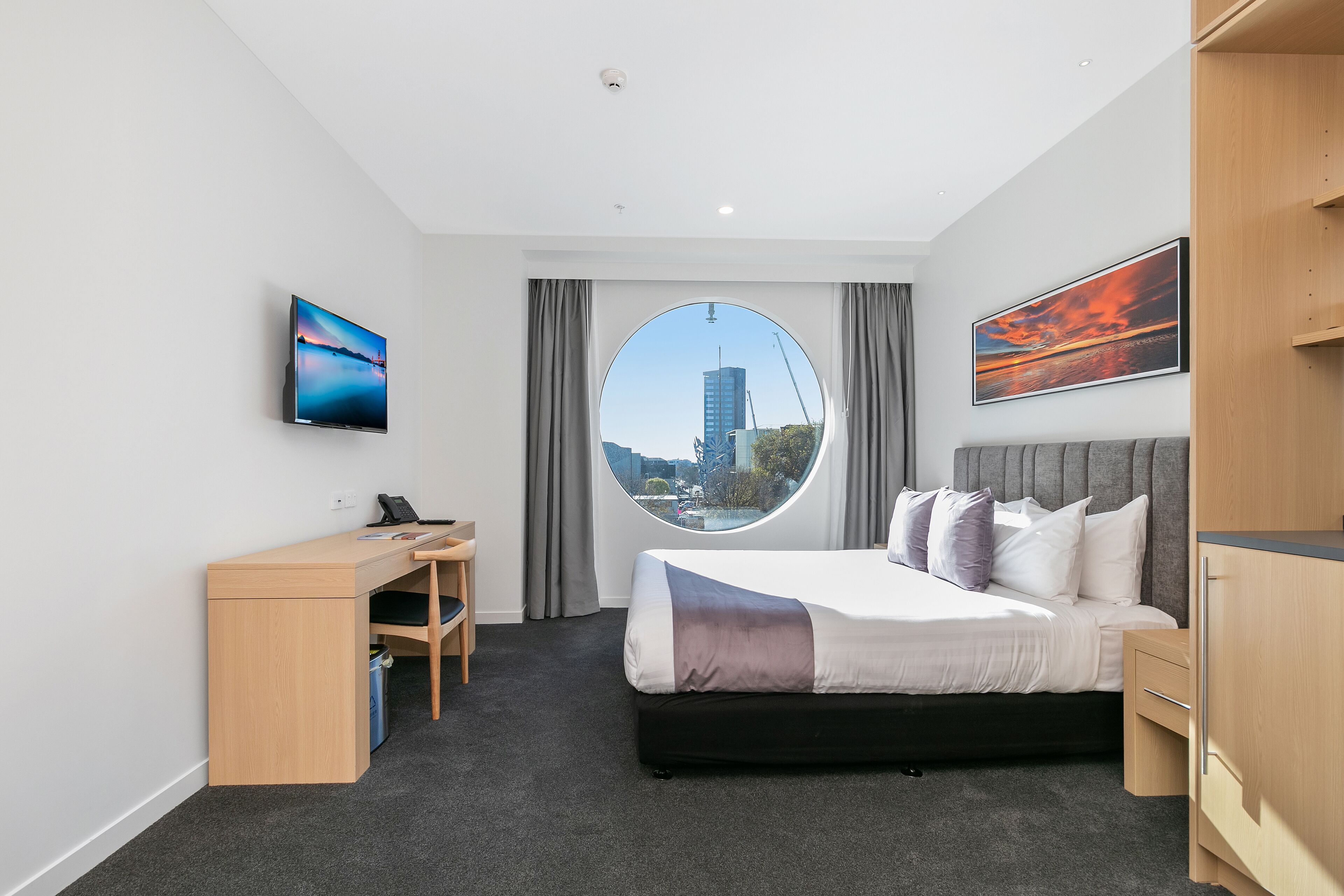 A modern hotel room with a large bed, a desk, a chair, a wardrobe, a round window with a city view, and a mounted TV.