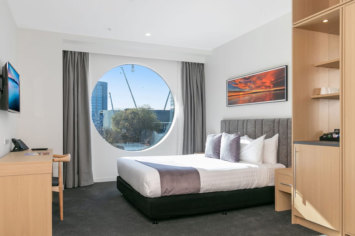 A modern hotel room with a large bed, a desk, a chair, a wardrobe, and a large window offering a view of the city.
