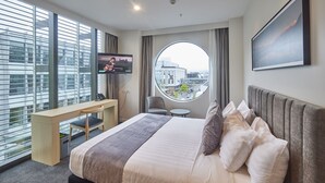 A modern hotel room with a large bed, a desk, a chair, a round table, a TV, and a window with a city view. - Christchurch City Hotel (Christchurch)
