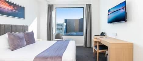 A hotel room with a large bed, a desk, a chair, and a view of a cityscape through a large window.