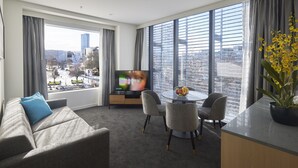 A modern hotel room with a large window, a flat-screen TV, a sofa with a blue pillow, a dining table with chairs, and a potted plant. - Christchurch City Hotel (Christchurch)