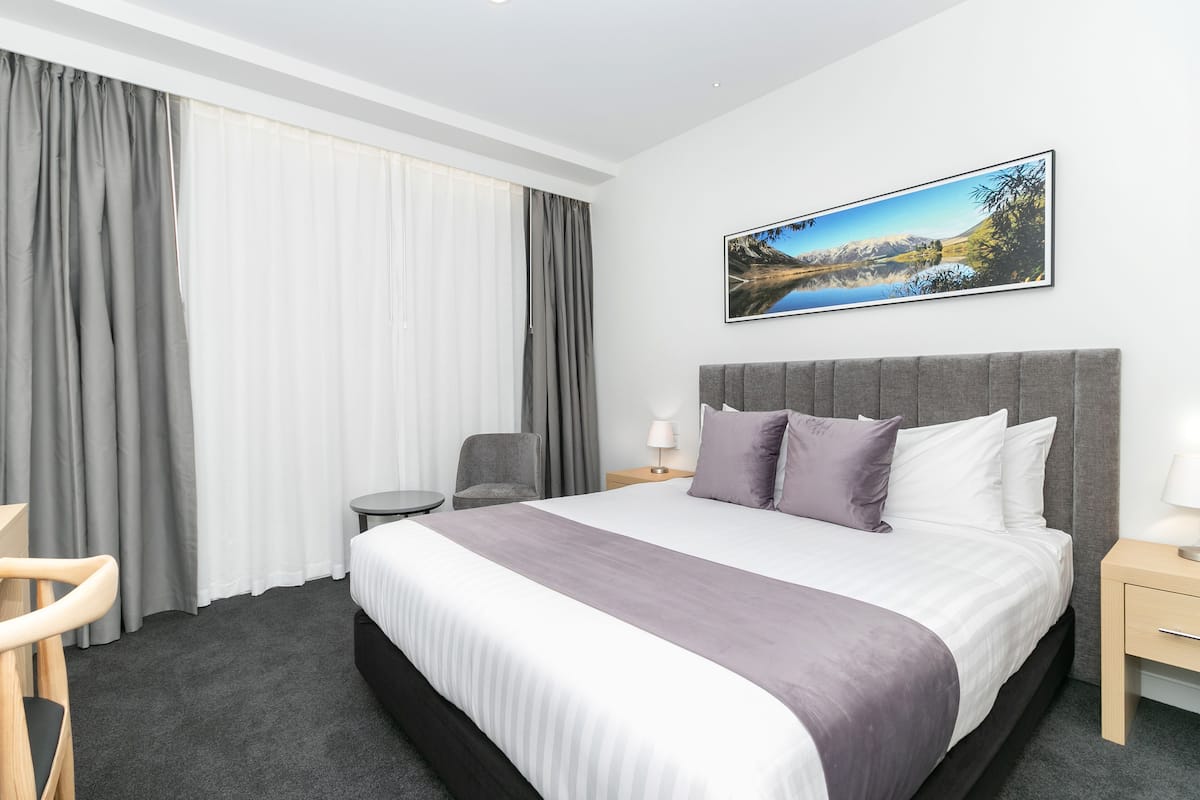 A hotel room with a large bed, a chair, a small table, and a painting on the wall.