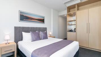 A hotel room with a large bed, a grey headboard, a nightstand with a lamp, a wardrobe, and a framed picture on the wall.