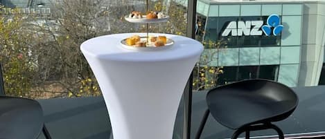 Una mesa blanca con una presentación de postres en una terraza en la azotea con vistas a la ciudad, dos sillas negras y un edificio con el rótulo 'ANZ' al fondo.