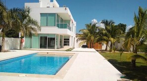 A modern two-story building with a swimming pool, surrounded by palm trees and a clear sky.