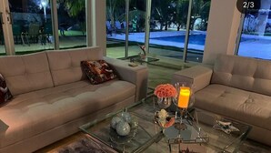 Uma sala de estar moderna com dois sofás voltados para uma mesa de centro de vidro. A sala possui janelas amplas com vista para uma piscina e área de estar externa.