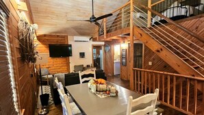A dining area with a long table and white chairs, a staircase, and a television mounted on the wall. - Aunt B`s Cabin  W/  Hot Tub!  Near Table Rock Lake (Lampe)