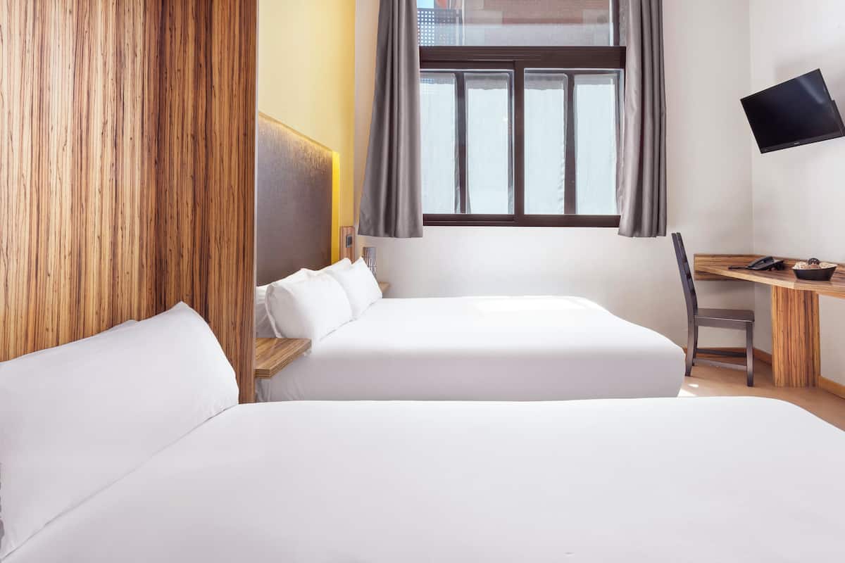 A hotel room with two beds, a desk, a chair, a TV mounted on the wall, and a window with curtains.
