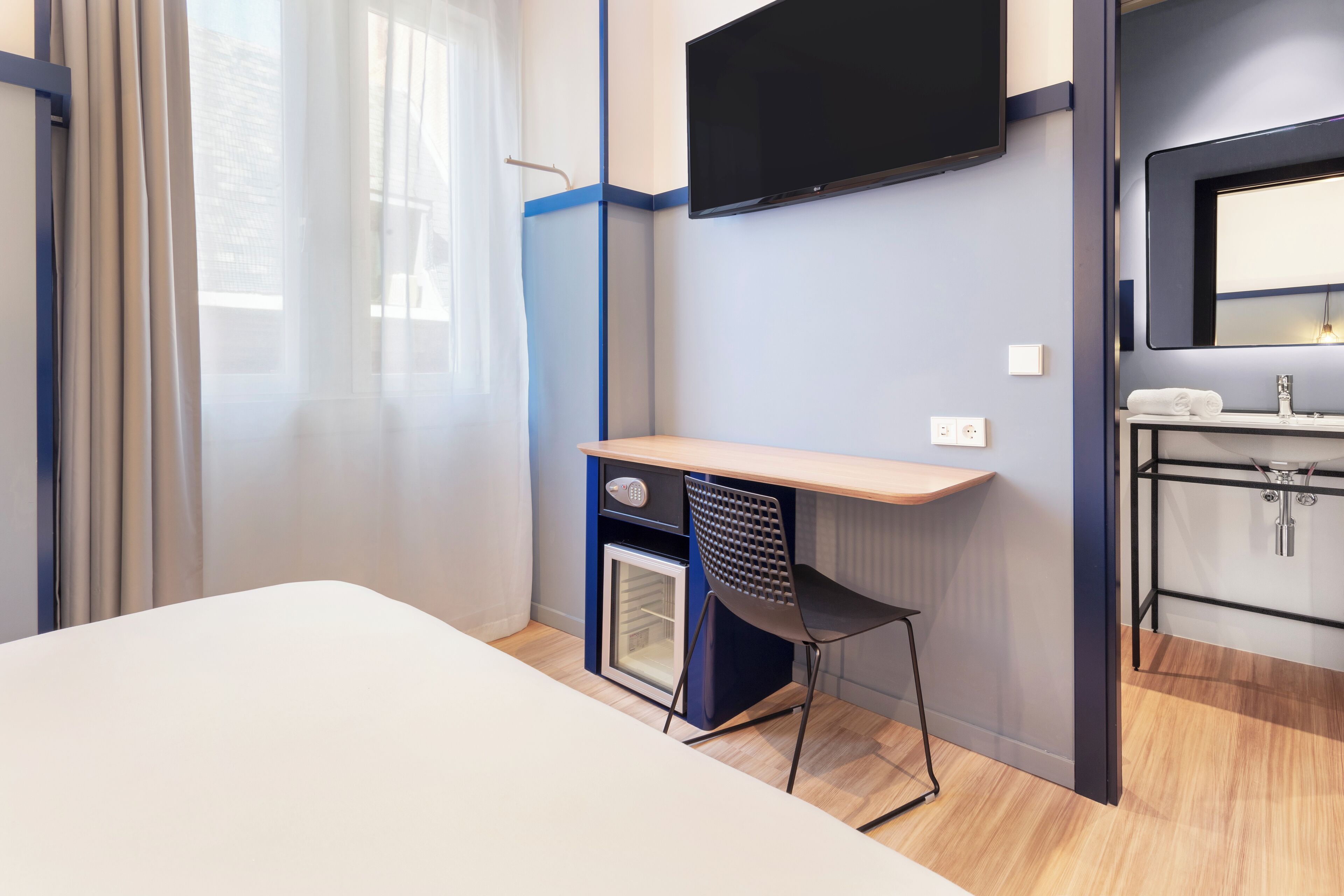 Una habitación de hotel con una cama, un escritorio y una silla, un televisor montado en la pared, una pequeña cocina y una ventana con cortinas.