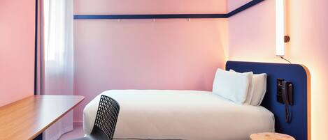 A compact hotel room with a single bed, a desk, a chair, and a small wooden stool.