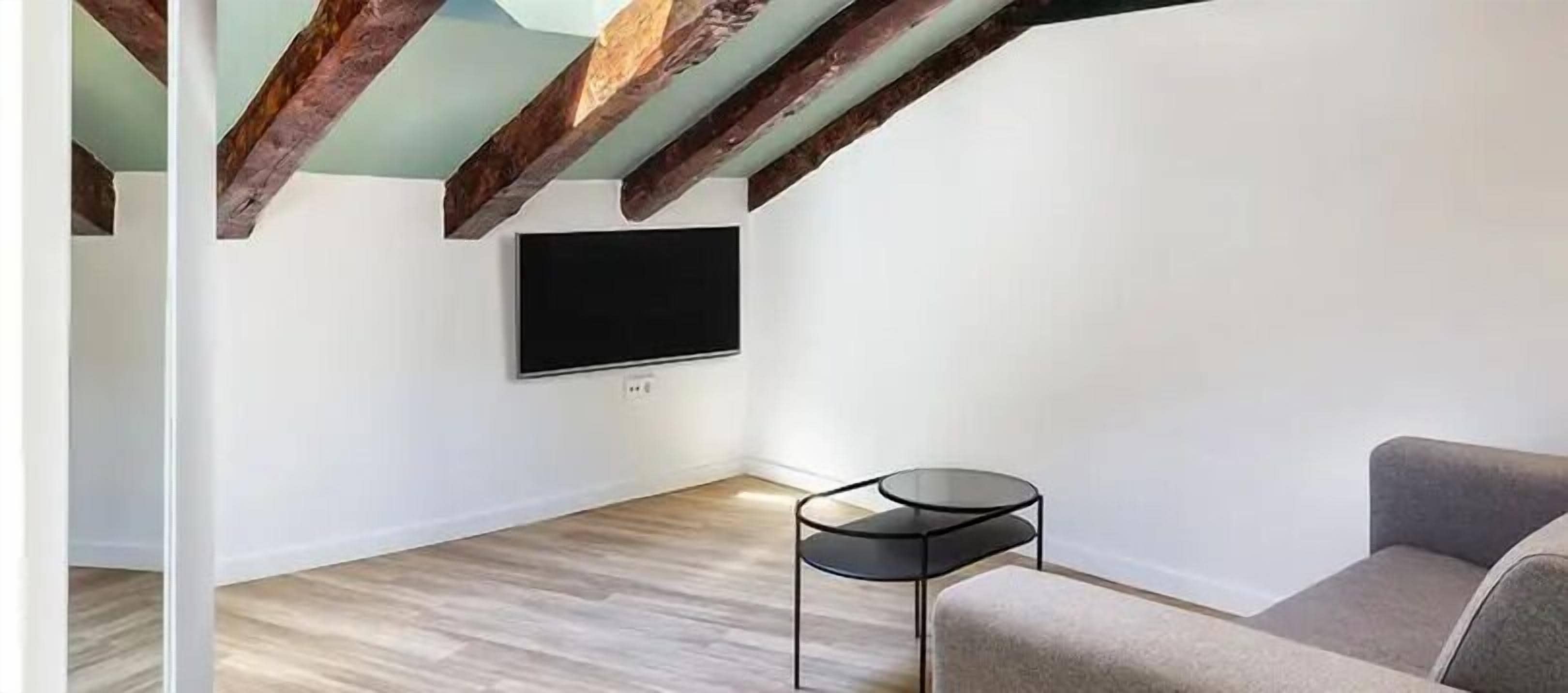 Un salon moderne avec des poutres apparentes, un téléviseur à écran plat fixé au mur, une petite table d’appoint ronde de couleur noire et un canapé gris.