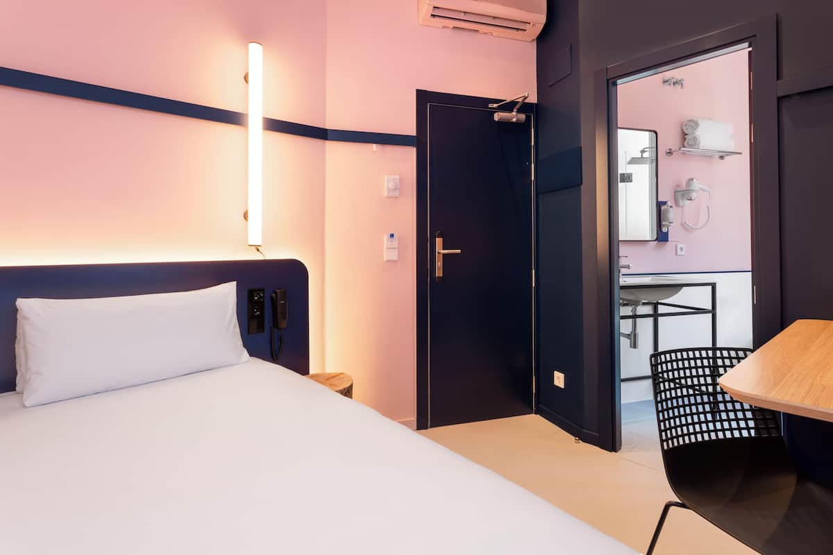A hotel room with a bed, bedside table, and a desk with a chair. There is a door leading to a bathroom with a mirror, towel rack, and sink.