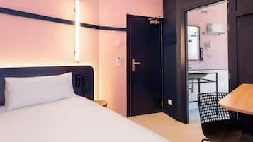 Habitación de hotel con cama, mesita de noche y escritorio con silla. Se ve una puerta que da a un baño con espejo, toallero y lavamanos.