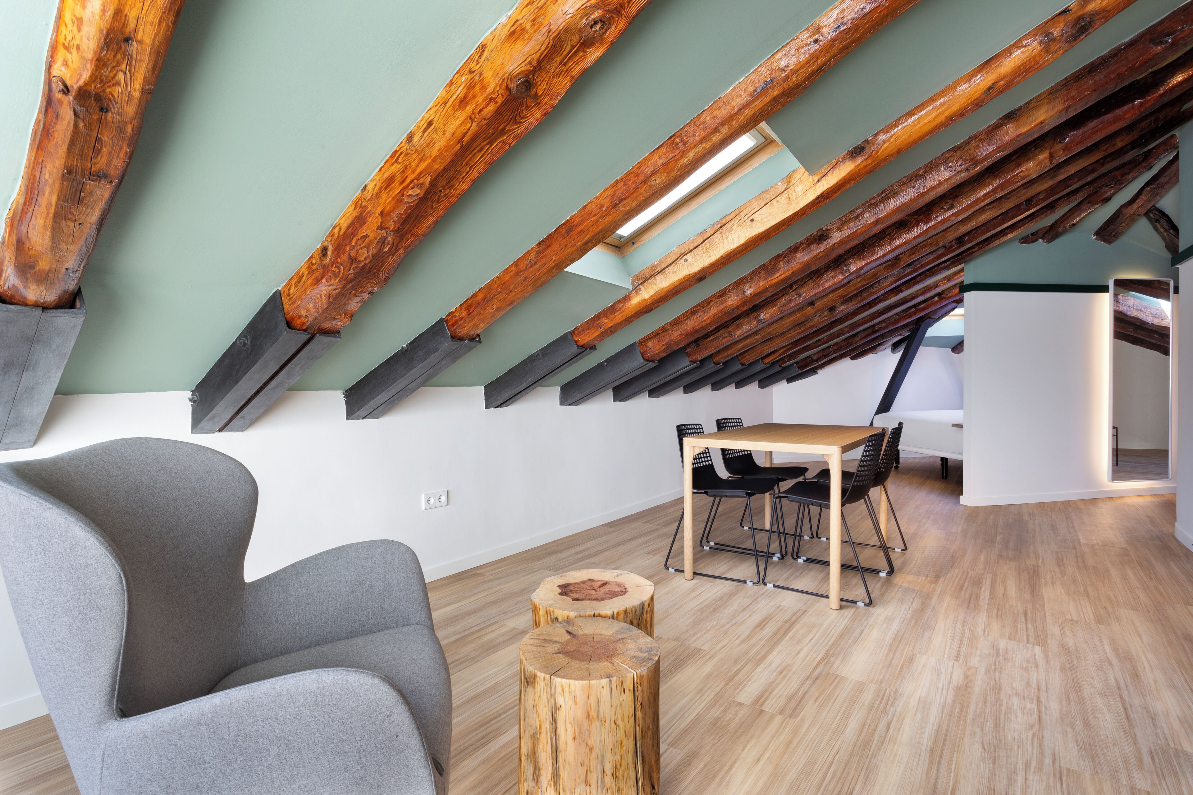 Un loft moderno con vigas de madera a la vista, un sillón gris, una mesa de centro de madera y un comedor con mesa blanca y sillas negras.