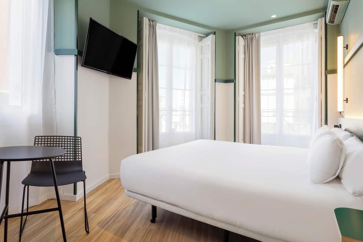 A hotel room with a bed, a TV mounted on the wall, and a chair.
