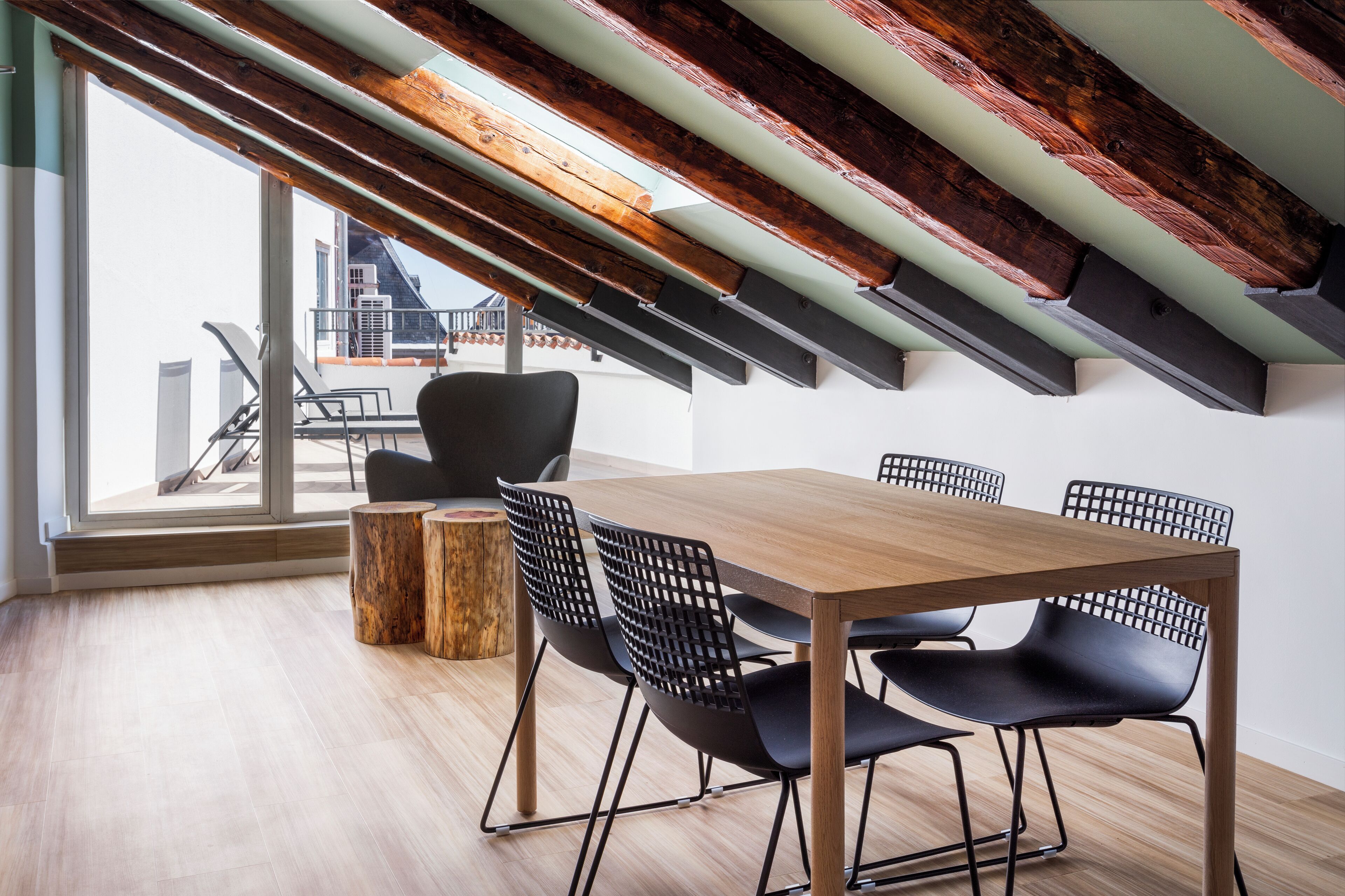 Un comedor moderno con una mesa de madera y sillas negras, un taburete de madera, y vistas a un paisaje urbano a través de amplios ventanales.
